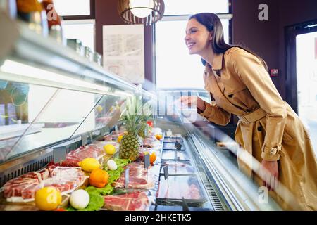 Glückliche junge Frau, die Fleisch aus dem Glasschrank im Lebensmittelgeschäft auswählt Stockfoto