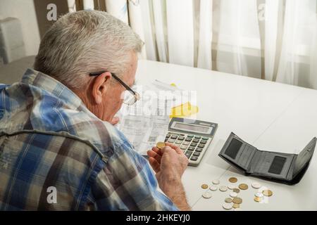 Älterer Mann, der am Tisch sitzt und die Finanzen berechnet. Alter Mann, der Rechnungen überprüft. Mann, der Münzen auf dem Tisch zählt. Pensionsberechnungskonzept Stockfoto