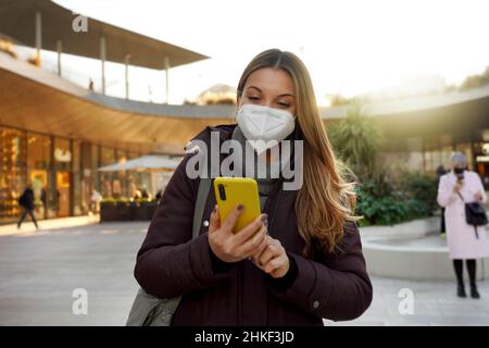 Porträt einer schönen Frau, die eine medizinische Gesichtsmaske trägt und das Internet WiFi am Telefon in der Stadt bei Sonnenuntergang nutzt Stockfoto