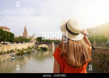 Besuch In Murcia, Spanien. Rückansicht eines touristischen Mädchens, das die Stadtlandschaft von Murcia mit der Puente Viejo Brücke und dem Glockenturm der Kathedrale, Spanien, genießt. Stockfoto