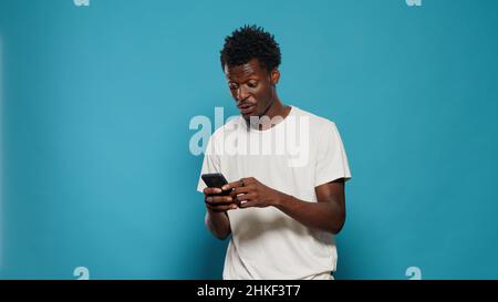 Lässige Person, die Smartphone anschaut und im Studio tanzt. Fröhlicher Mann, der Musik und Lieder hört, während er das Telefon mit Touchscreen in der Hand hält und über einem isolierten Hintergrund steht. Stockfoto