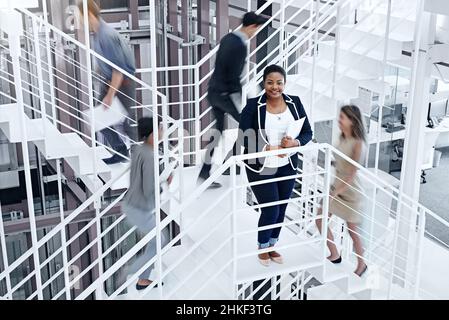 Damit beschäftigt, Erfolg zu erreichen. Porträt einer jungen professionellen Person, die auf einer Treppe steht, mit Kollegen, die um sie herum eilen. Stockfoto