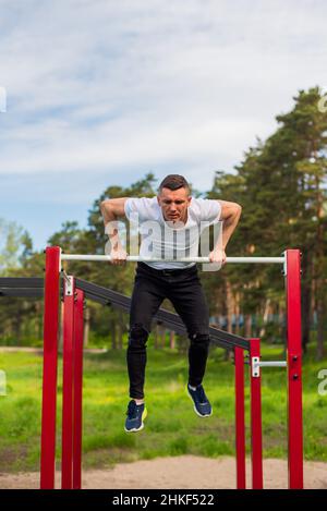 Der kaukasische Mann zieht sich auf einer horizontalen Stange auf einem Sportplatz hoch. Stockfoto