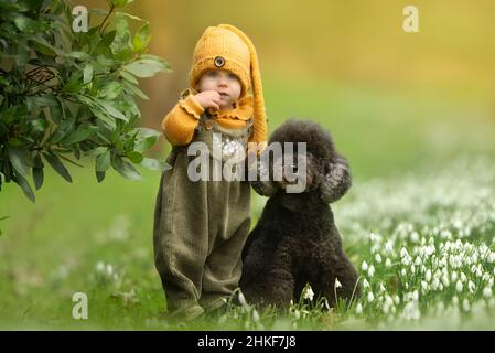 Kleine Mädchen mit schwarzem Pudelhund in lässiger Kleidung, die im Februar im Freien in Schneeglöckchen stehen Stockfoto