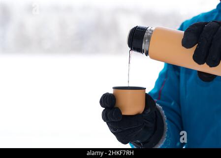 Weibliche Hand gießt heißen Tee oder Kaffee in einen Becher aus einer Thermoskanne im Winterwald. Wärmendes Getränk bei kaltem Wetter. Stockfoto