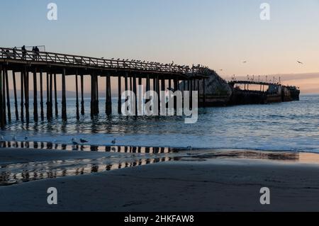 Ein wunderschöner Sonnenuntergang über dem Strand in der Nähe von Aptos, Kalifornien, der den alten, verödnisten Pier und ein altes Schiffswrack hervorhebt. Stockfoto