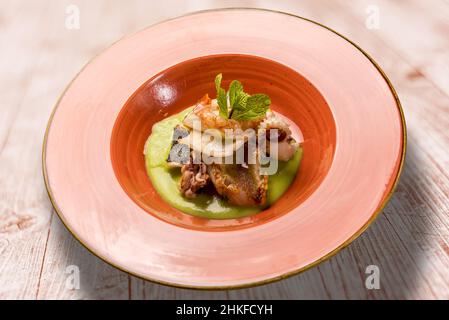 Fischeintopf auf Erbsencreme - Garnelen, Steinbutt, Tintenfisch und Tintenfische in Steingutform auf Holztisch Stockfoto