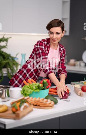 Schöne Frau mit kurzen Haaren und rotem Hemd macht frische Lebensmittel zu Hause. Gesundes Lifestyle-Konzept Porträt einer schönen jungen Frau, die zu Hause in der Küche ein hausgemachtes Essen zubereitet. Stockfoto