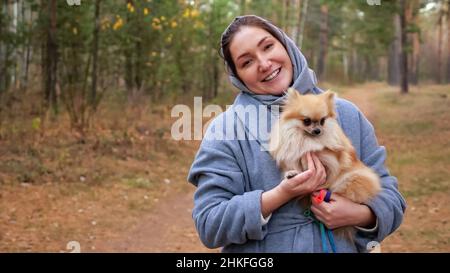Junge Frau, die den pommerschen Mini-spitz in den Armen hält, während sie im Herbstpark spazieren geht Stockfoto