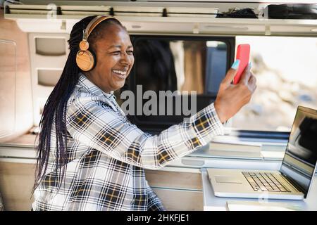 Afrikanische ältere Frau, die Spaß mit Videoanrufen und das Tragen von Kopfhörern im Camper Mini-Van - Fokus auf Gesicht Stockfoto