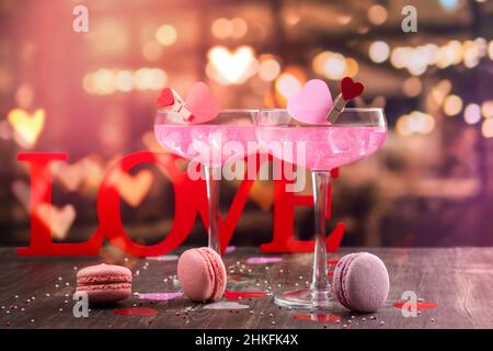 Zwei Gläser mit rosafarbenem Champagner, Makronen-Keksen und Holzwort LOVE auf einer Theke. Feier Valentinstag, Grußkarte. Speicherplatz kopieren Stockfoto