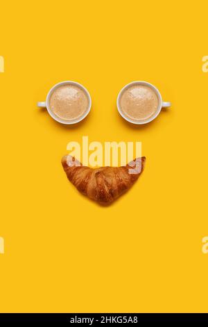 Zwei Tassen Kaffee mit Schaumstoff und einem Croissant bilden ein lächelndes, fröhliches Gesicht auf einem gelben Hintergrund mit Platz für Text und Text Stockfoto