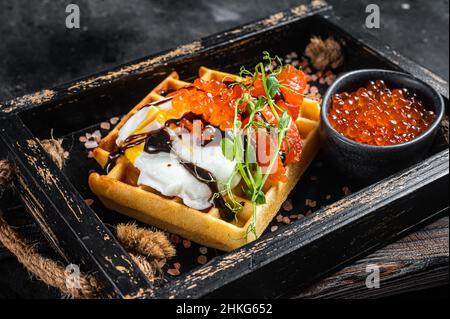 Köstliche Waffeln mit geräuchertem Lachsfilet, pochiertem Ei und rotem Kaviar. Schwarzer Hintergrund. Draufsicht Stockfoto