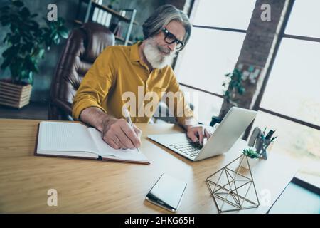 Foto von hübscher reifer Kerl gekleidet gelbes Hemd Schreiben Buch Eingabe moderne Gadget drinnen Werkstatt-Workstation Stockfoto