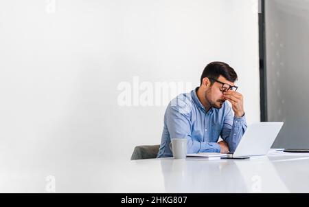 Büromitarbeiter mit schmerzhaften Kopfschmerzen bei der Arbeit am Schreibtisch und ganztägig sitzend mit einem Laptop oder Notebook, die Kopfschmerzen bei kranker Arbeit haben Stockfoto