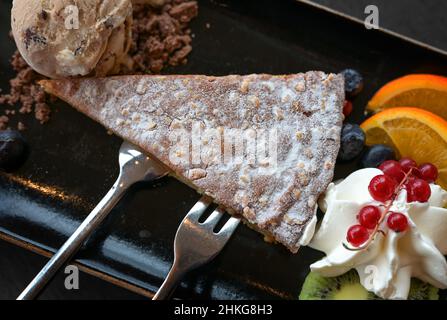 Dessert aus hausgemachter Mandelkuchen mit Eis und frischen Früchten auf schwarzem Teller mit Besteck, Blick von oben Stockfoto