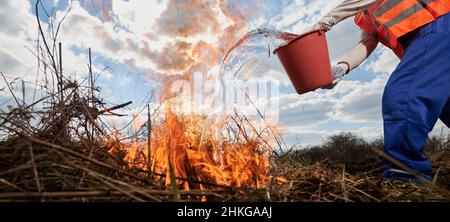 Feuerwehrmann-Ökologe löscht Feuer auf dem Feld mit bewölktem Himmel auf dem Hintergrund. Nahaufnahme eines Mannes, der Eimer hält und Wasser auf brennendes trockenes Gras gießt. Naturkatastrophen und ökologische Demage Konzept. Stockfoto