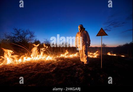 Feuerwehrmann, der nachts auf dem Feld mit einem Wildfeuer arbeitet. Mann in Anzug und Gasmaske neben brennendem Gras mit Rauch und gelbem Dreieck mit Totenkopf und Kreuzknochen Warnschild. Konzept für Naturkatastrophen. Stockfoto