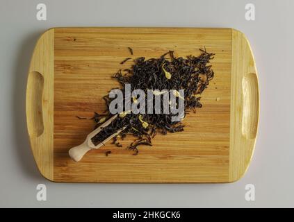 Der Tee ist bereit zum Brühen. Spachtel zum Brühen von Tee. Hintergrund von duftenden schwarzen Teeblättern mit getrockneten Jasminblüten. Tee auf einem Holzbrett. Stockfoto