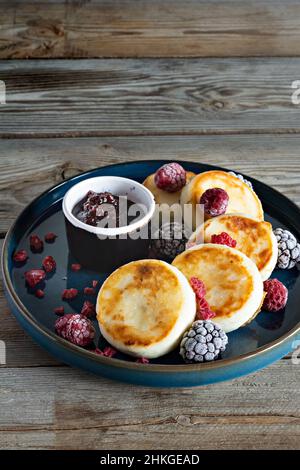 Frischer Hüttenkäse oder Ricotta-Pfannkuchen (Syrniki, Krapfen) auf einem blauen Teller mit frischen Beeren und Marmelade. Leckeres gesundes Hüttenkäse-Frühstück. Stockfoto