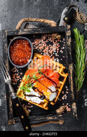 Köstliche Waffeln mit geräuchertem Lachsfilet, pochiertem Ei und rotem Kaviar. Schwarzer Hintergrund. Draufsicht Stockfoto