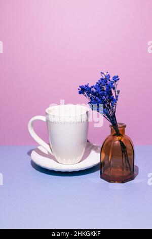 Weiße Kaffeetasse mit Vase aus blauen trockenen Blumen auf blauem Tisch. Rosa Wandhintergrund Stockfoto