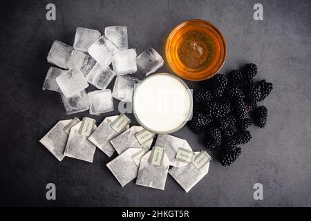 Eisted Blackberry Infused Earl Grey Tea Zutaten: Overhead-Ansicht von Teebeuteln, Milchpulver, Honig und Brombeeren mit Eiswürfeln Stockfoto