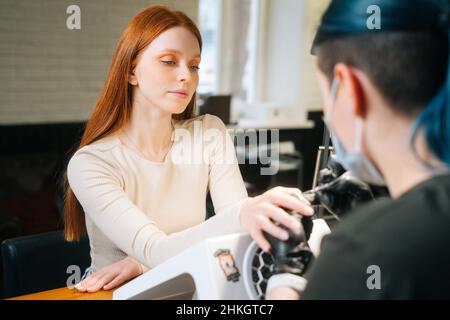 Nahaufnahme von der Rückseite einer nicht erkennbaren professionellen Manikuristin, die im Schönheitssalon mit einem speziellen Werkzeug die Nagelhaut hebt, um eine attraktive junge Frau zu sehen. Stockfoto