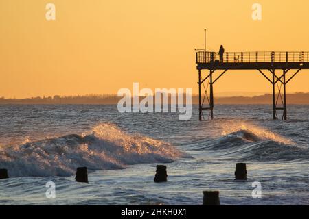 Ein eingeflügelter Fischer mit Angelrute, der am Ende des Bognor Regis Piers bei Sonnenuntergang steht und Wellen am Ufer brechen. Stockfoto