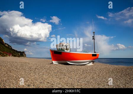 Einzelnes rotes kleines Fischerboot am Kiesstrand. E293 rotes Fischerboot am Strand von Branscome Dorset. Ruhiges Meer im Hintergrund mit blauem Himmel und Wolken mit Stockfoto