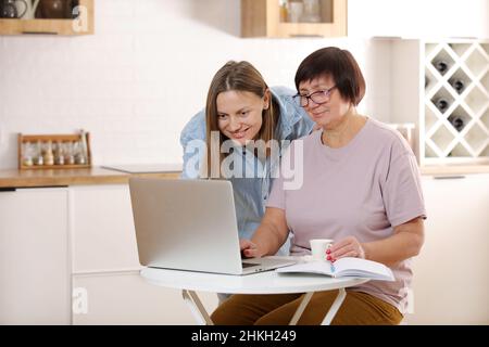 Junge Frau, die ältere Mutter lehrt, das Internet auf dem Laptop zu Hause zu nutzen. Tochter hilft ihrer älteren Mutter, es online mit ihrem persönlichen Konto herauszufinden Stockfoto