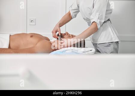 Masseur arbeitet mit dem Kinn des bärtigen Klienten im Klinikbüro Stockfoto