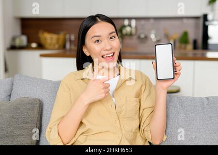 Glückliche junge asiatische Frau in eleganter Freizeitkleidung freut sich über den Erfolg und demonstriert dies auf einem Smartphone-Bildschirm, während sie auf dem Sofa sitzt Stockfoto