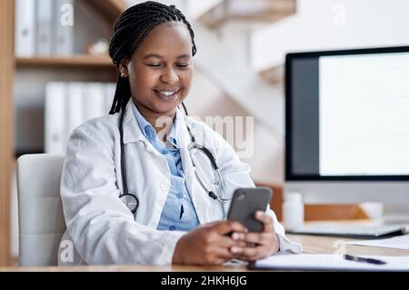 Sie begrüßt Texte ihrer Patienten. Aufnahme einer jungen Ärztin, die ein Mobiltelefon benutzte, während sie in ihrem Büro arbeitete. Stockfoto