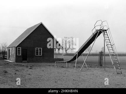 1950s, historischer, äußerer, Seitenansicht eines traditionellen Sportpavillons aus der Zeit, der mit einem Holzrahmen auf einem Ziegelsockel und einem hoch abfallenden Dach gebaut wurde. Architektonisch unverwechselbar mit einer überdachten Veranda oder Veranda an der Vorderseite, wurden diese Arten von einstöckigen Gebäuden am Rande von Sportplätzen zu dieser Zeit auf vielen Freizeit- und Sportplätzen in Großbritannien gesehen, die Umkleidekabinen für Mannschaftssportarten wie Cricket, Rugby und Fußball zur Verfügung stellten. Neben diesem neu errichteten Pavillon in Witney, Oxford, England, Großbritannien, gibt es einen Spielplatz für Kinder mit Medaillenrutsche. Stockfoto