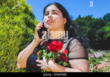 Porträt einer jungen kaukasischen lateinamerikanischen Frau in der Liebe dramatisch in einem öffentlichen Park mit Rosen und Bäumen, steht sie am Telefon und umarmt Stockfoto