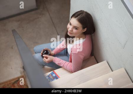 Draufsicht auf eine schöne junge Frau, die sich entspannt auf der Treppe in ihrer modernen Wohnung sitzt und Tee trinkt Stockfoto