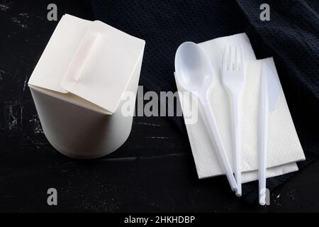 Lebensmittelverpackungen aus Pappe und Plastiklöffel, Messer und Gabel auf schwarzem Hintergrund. Take-away-Box und Einwegbesteck. Papierbehälter für Nudeln. Lu Stockfoto