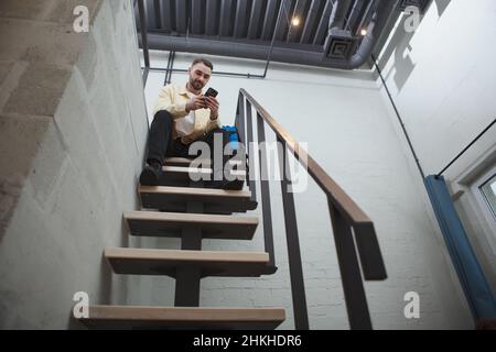 Aus dem niedrigen Winkel aufgenommen, in dem ein gutaussehender Mann mit einem Smartphone sitzt, der mit seinem Rucksack auf der Treppe im modernen Hotelzimmer sitzt und Platz zum Kopieren verwendet Stockfoto