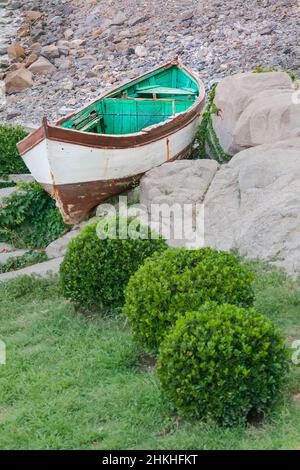 Ein Holzboot liegt an der Küste zwischen großen Steinen und grünen Büschen Stockfoto