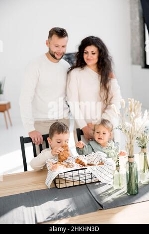 Fröhliche Familie mit kleinen Sohn und Tochter, die ein Frühstück haben und die Wochenenden zu Hause verbringen. Schöne glückliche Eltern stehen in der Küche und lächeln Stockfoto