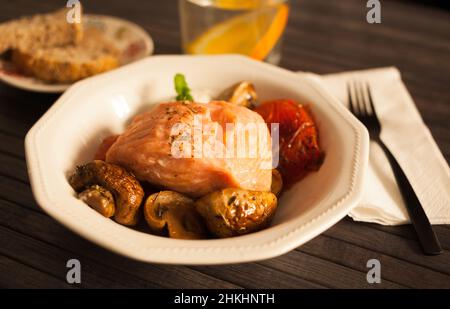 Gedünstetes Lachsfilet auf einem Kissen aus gebratenen Pilzen und Tomaten in einer weißen Schüssel Stockfoto
