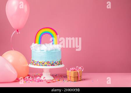 Blauer Geburtstagstorte mit Zuckerwatte und einem bunten Regenbogen auf einem rosa Hintergrund, umgeben von Partyballons, Konfetti-Streuseln und einem Geschenk. Stockfoto