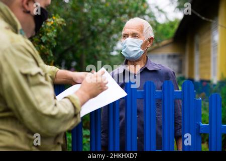 Älterer Mann in Schutzmaske, der mit dem Vertreter der Versicherungsgesellschaft kommuniziert, während er am Zaun seines Landhauses steht Stockfoto