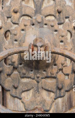 Nahaufnahme einer alten hölzernen verzierten Tür mit geschnitztem Holz mit korrodierten und rostigen Beschlägen, Elementen eines Türklopfers in Form eines menschlichen Kopfes. Stockfoto