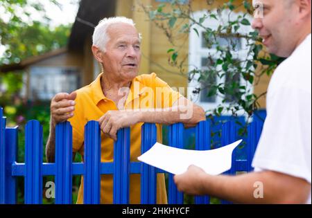 Leitender Mann, der mit dem Vertreter der Versicherungsgesellschaft kommuniziert, während er am Zaun seines Landhauses steht Stockfoto