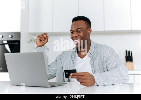 Internet-Shopping. Attraktiv, zufrieden, jung, erwachsen, afroamerikanischer Kerl, Sitzen an einem Tisch in der Küche, Online-Einkäufe auf Websites und Anwendungen, Eingabe von Kartendaten, lächelnd. Stockfoto