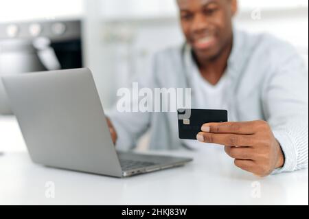 Online-Shopping im Internet. Unverschämter schwarzer Mann kauft von zu Hause aus in Online-Shops, stöbert mit einem Laptop in Anwendungen auf Websites und hält die Kreditkarte in der Hand, lächelt, gibt Informationen ein Stockfoto