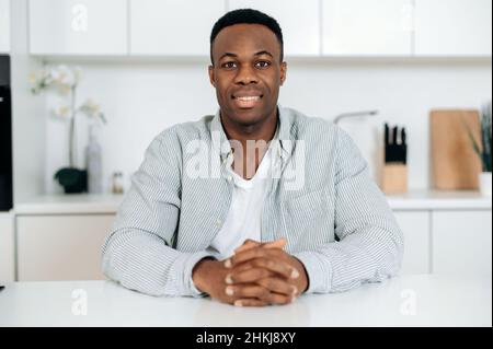 Porträt eines attraktiven, angenehmen afroamerikanischen Mannes, der in legerer Kleidung gekleidet ist, zu Hause in der Küche am Tisch sitzt und freundlich lächelnd direkt auf die Kamera blickt Stockfoto