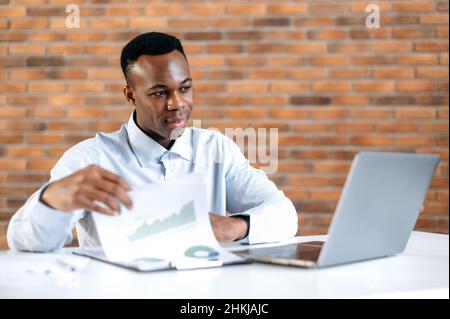 Fokussierter afroamerikanischer Geschäftsmann, der Finanzdiagramme, Gewinne und am Arbeitsplatz sitzt. Smart schwarz männlich Student studiert online, hört Online-Vorlesung, Freiberufler ist upskills, mit Laptop Stockfoto